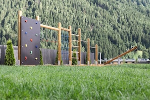 Un parque infantil con juegos de escalada y columpios, rodeado de césped y una colina boscosa al fondo.