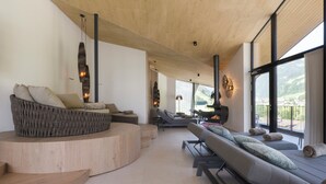 Un salon moderne avec un banc en bois aux lignes courbes, un canapé et un long banc agrémenté de coussins. De grandes fenêtres offrent une vue sur l’extérieur.