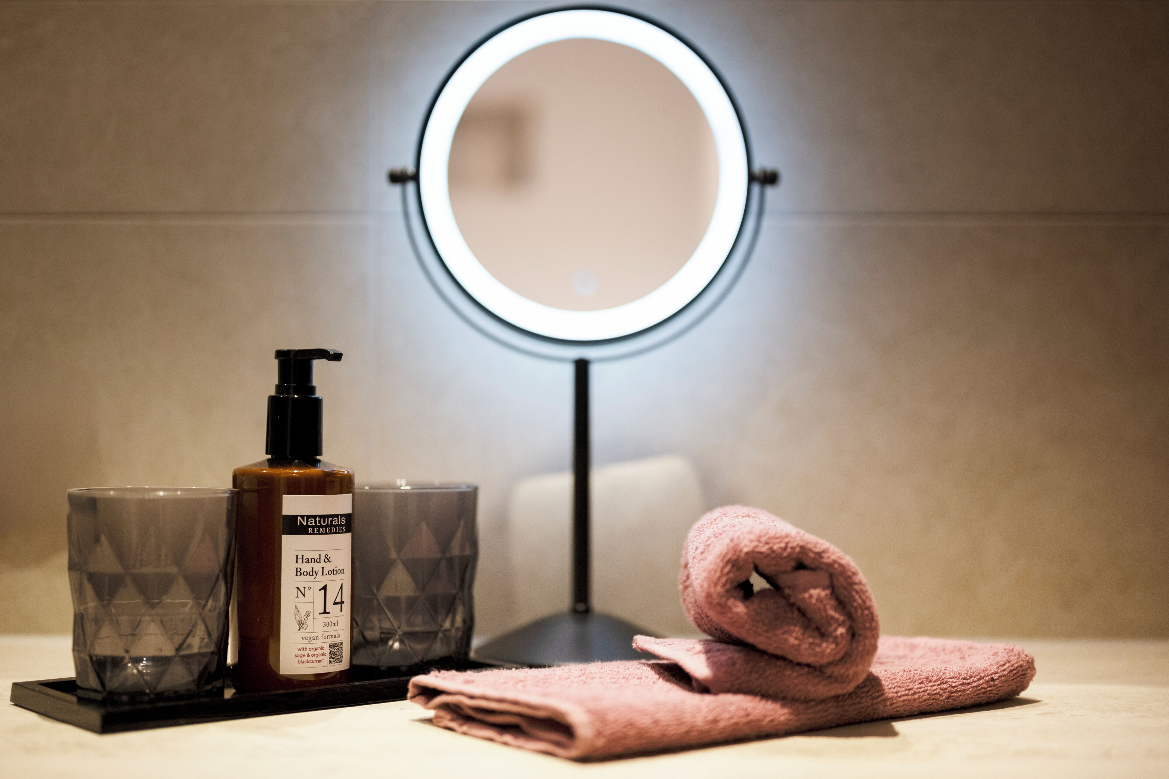 Un tocador de baño con un espejo redondo, una botella de crema corporal y una toalla doblada.