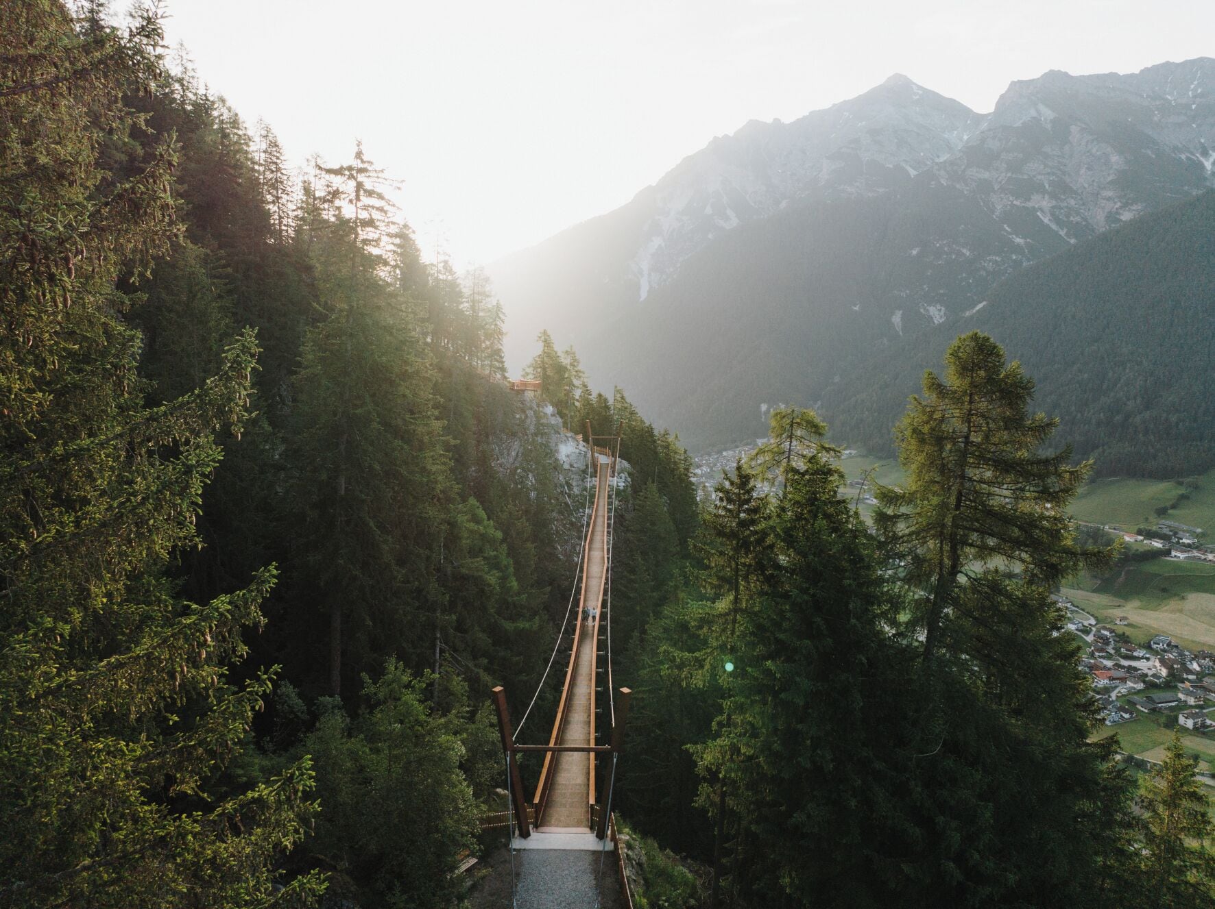A suspension bridge in a forested area with a view of a valley and mountains in the background.