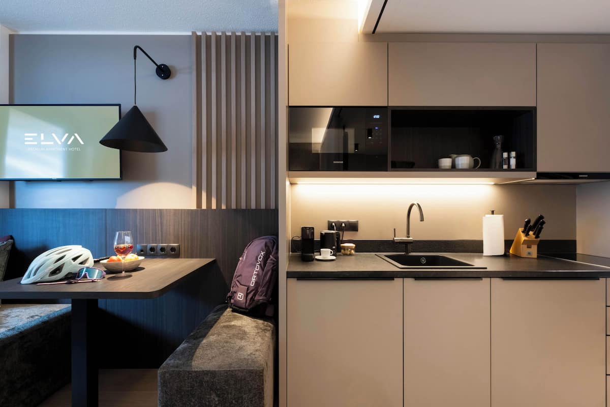 Una cocina moderna con horno empotrado, un lavamanos y una cafetera. Hay una zona de estar con un banco y una mesa.