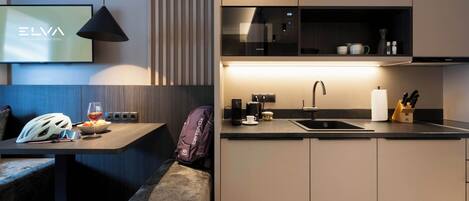 Una cocina moderna con horno empotrado, un lavamanos y una cafetera. Hay una zona de estar con un banco y una mesa.
