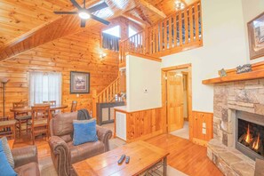 A cozy cabin interior with a wooden interior, a stone fireplace, a dining area, and a staircase leading to an upper level.