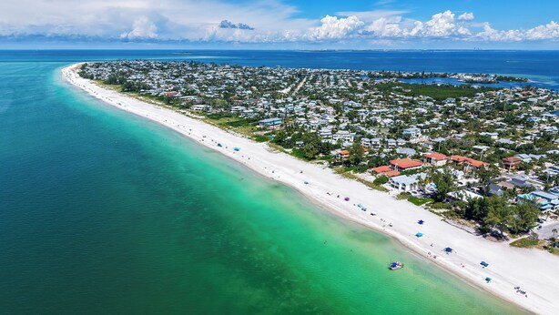 An aerial view of a coastal town with a long beach, clear turquoise waters, and a densely packed residential area with houses and buildings. - La Casa AMI Beachside-Beachfront Condo w/ Heated Pool, Hot tub & Garage Parking (Holmes Beach)