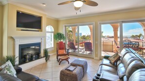 A living room with a fireplace, a large TV, a ceiling fan, and a view of the outdoors. - La Casa AMI Beachside-Beachfront Condo w/ Heated Pool, Hot tub & Garage Parking (Holmes Beach)