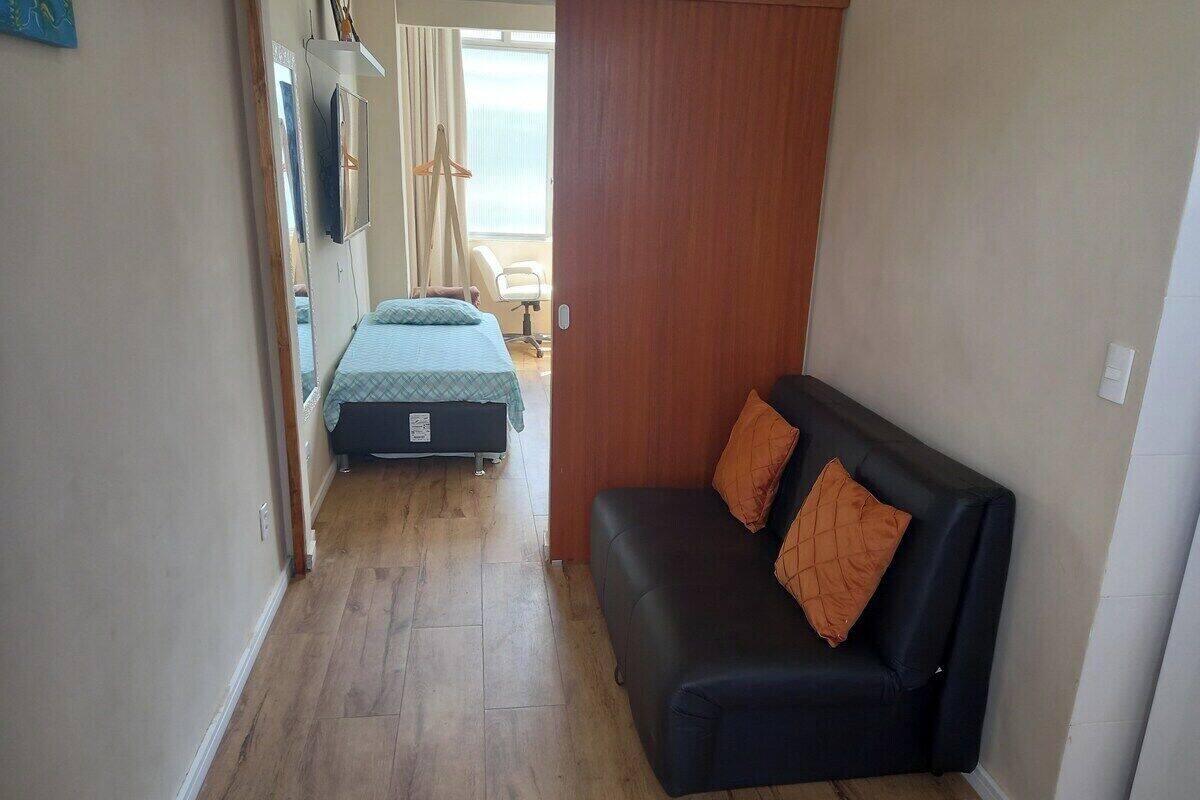 Una habitación compacta con un sofá negro, una cama con colcha azul y suelo de madera.