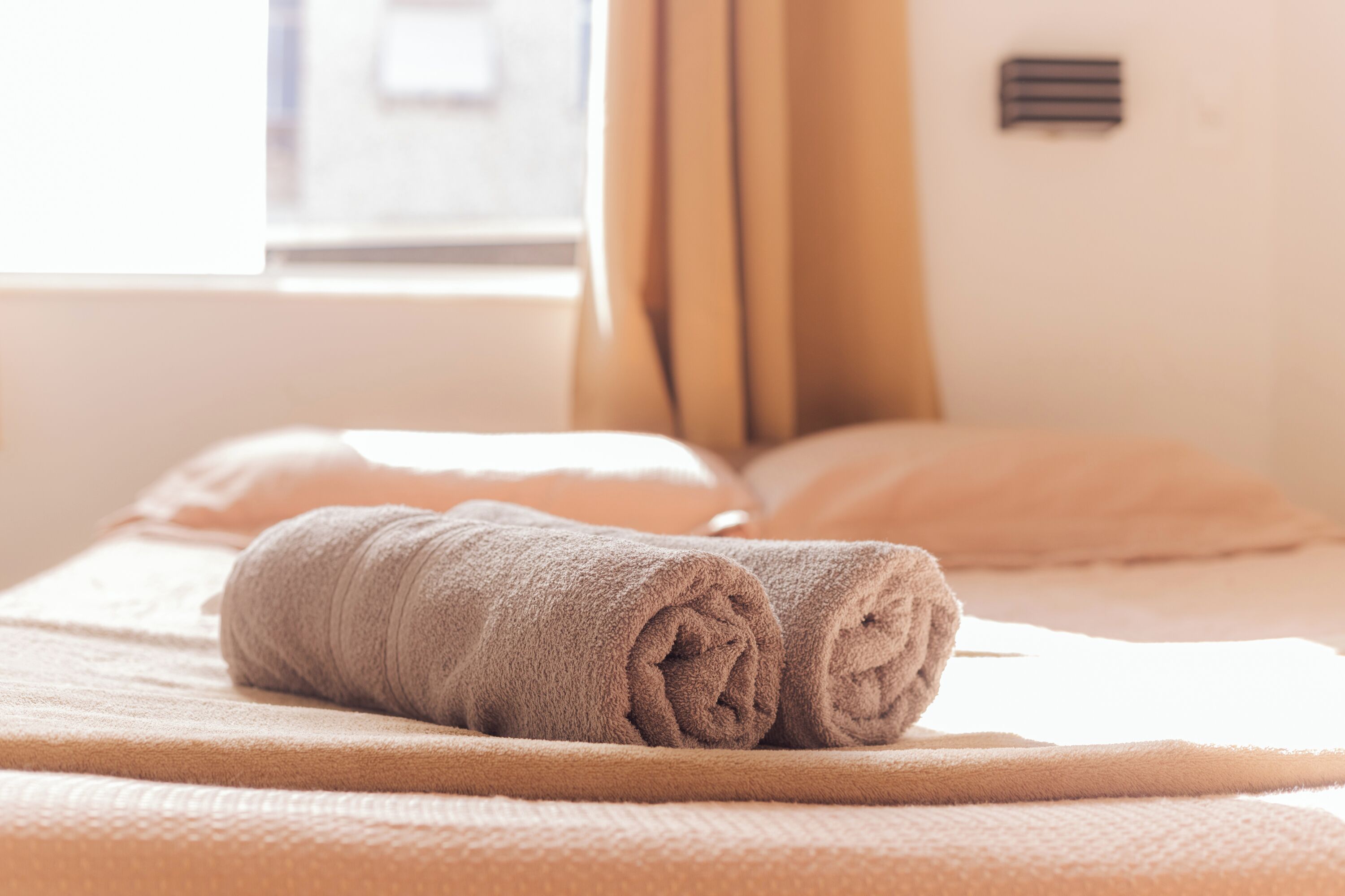 A neatly made bed with a rolled towel, a window with curtains, and a vent on the wall.