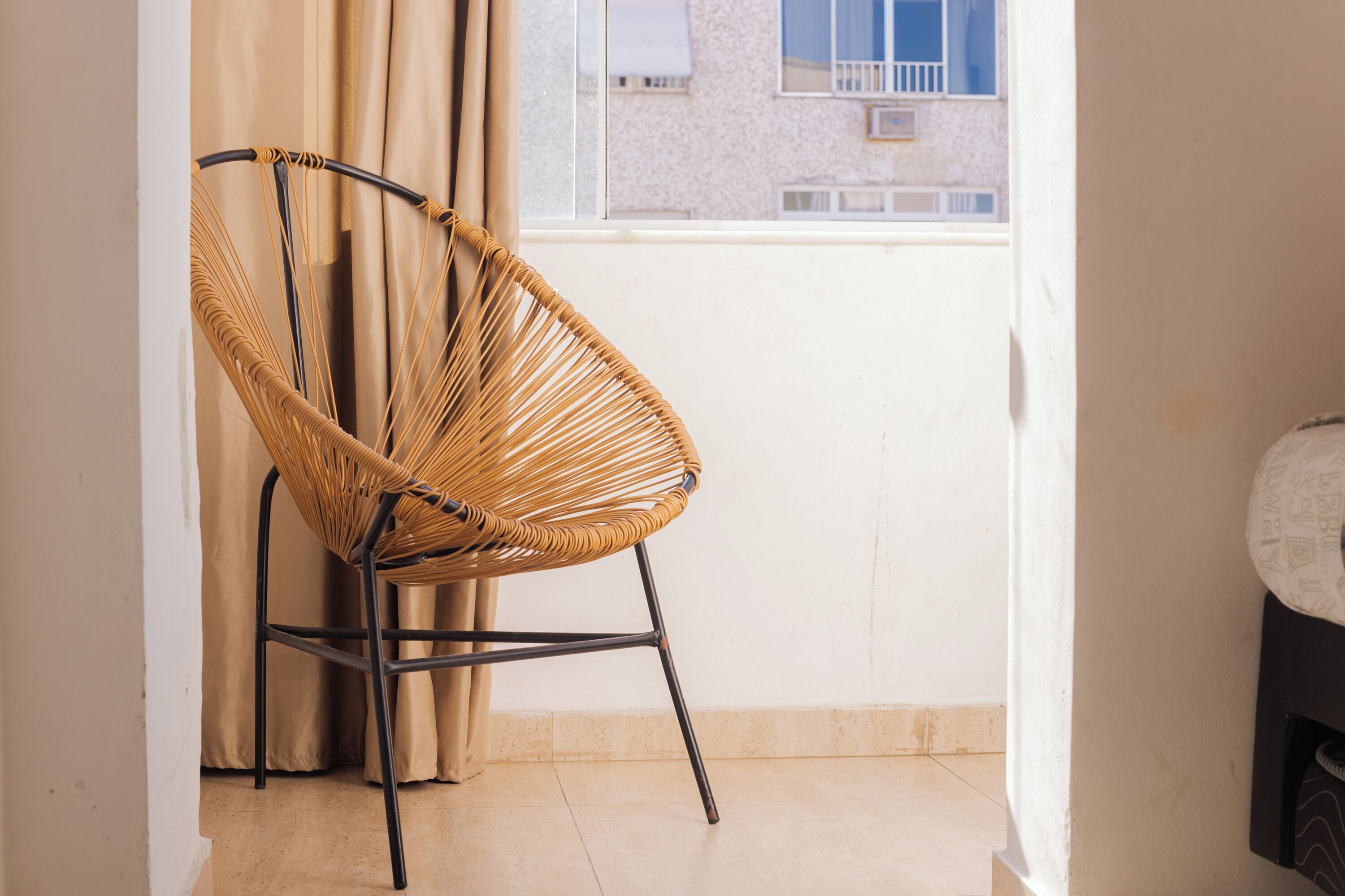 A wicker chair with metal legs in a room with a window, beige curtains, and a tiled floor.