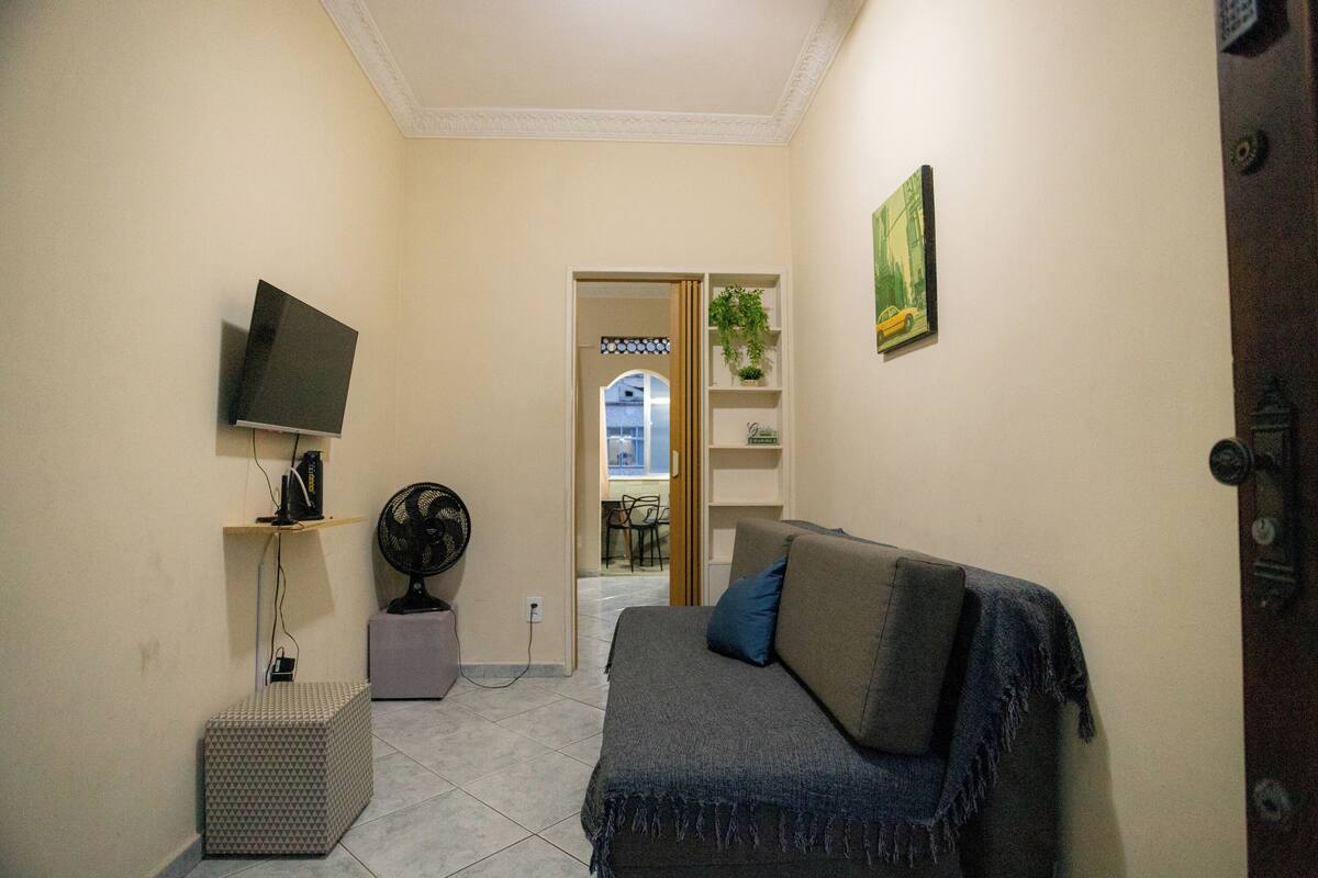 Una habitación pequeña y bien iluminada con un sofá gris, un televisor de pantalla plana montado en la pared, un pequeño ventilador y piso de baldosas.