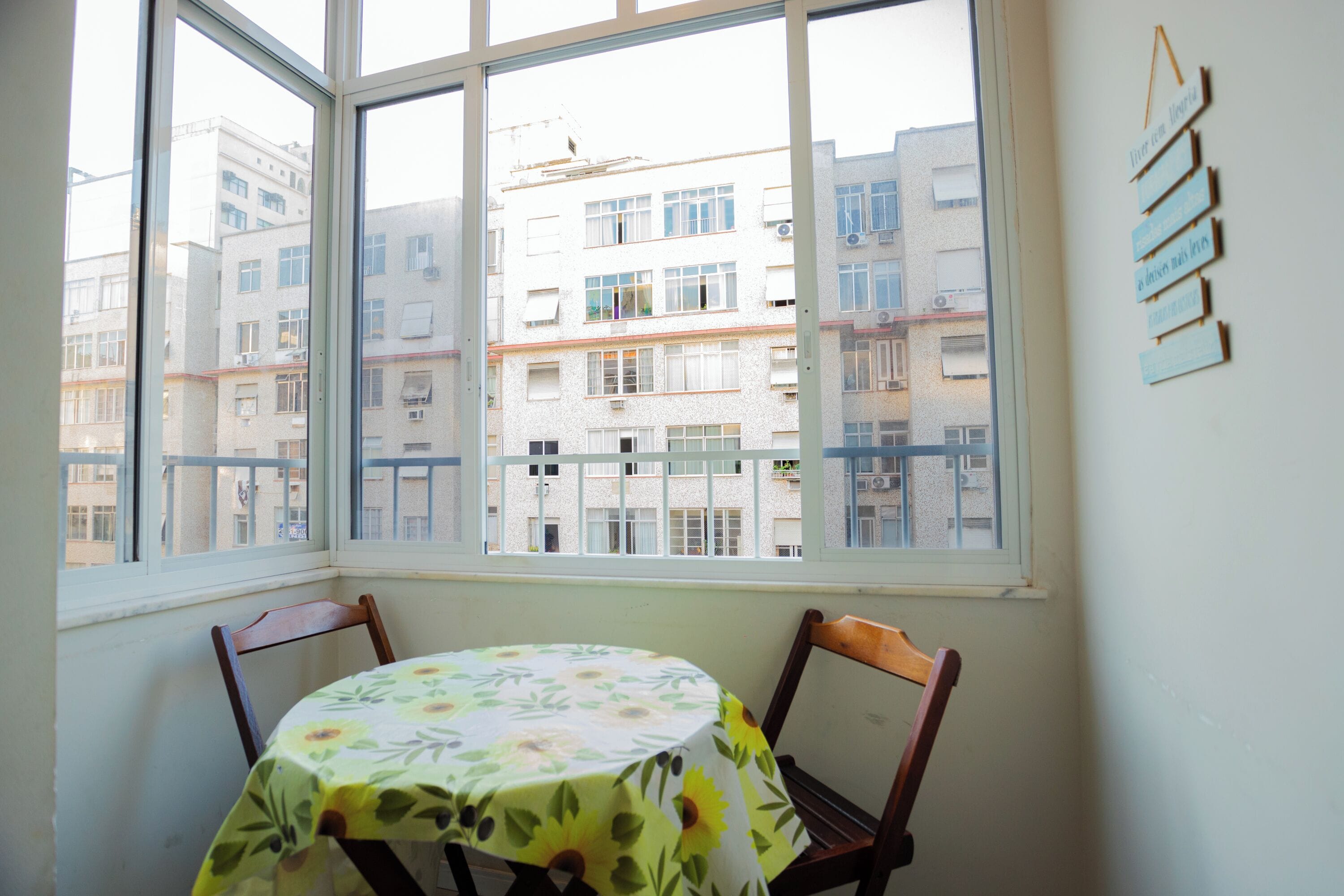 Une pièce comprenant une table et deux chaises, une fenêtre donnant sur des immeubles et un mur sur lequel est accrochée une enseigne.