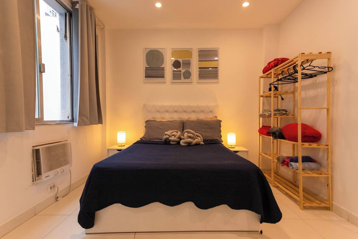 Una cama bien tendida con una colcha azul oscuro, un cabecero capitonado y dos lámparas de noche. Se observa una estantería de madera con ropa y un radiador en la pared.