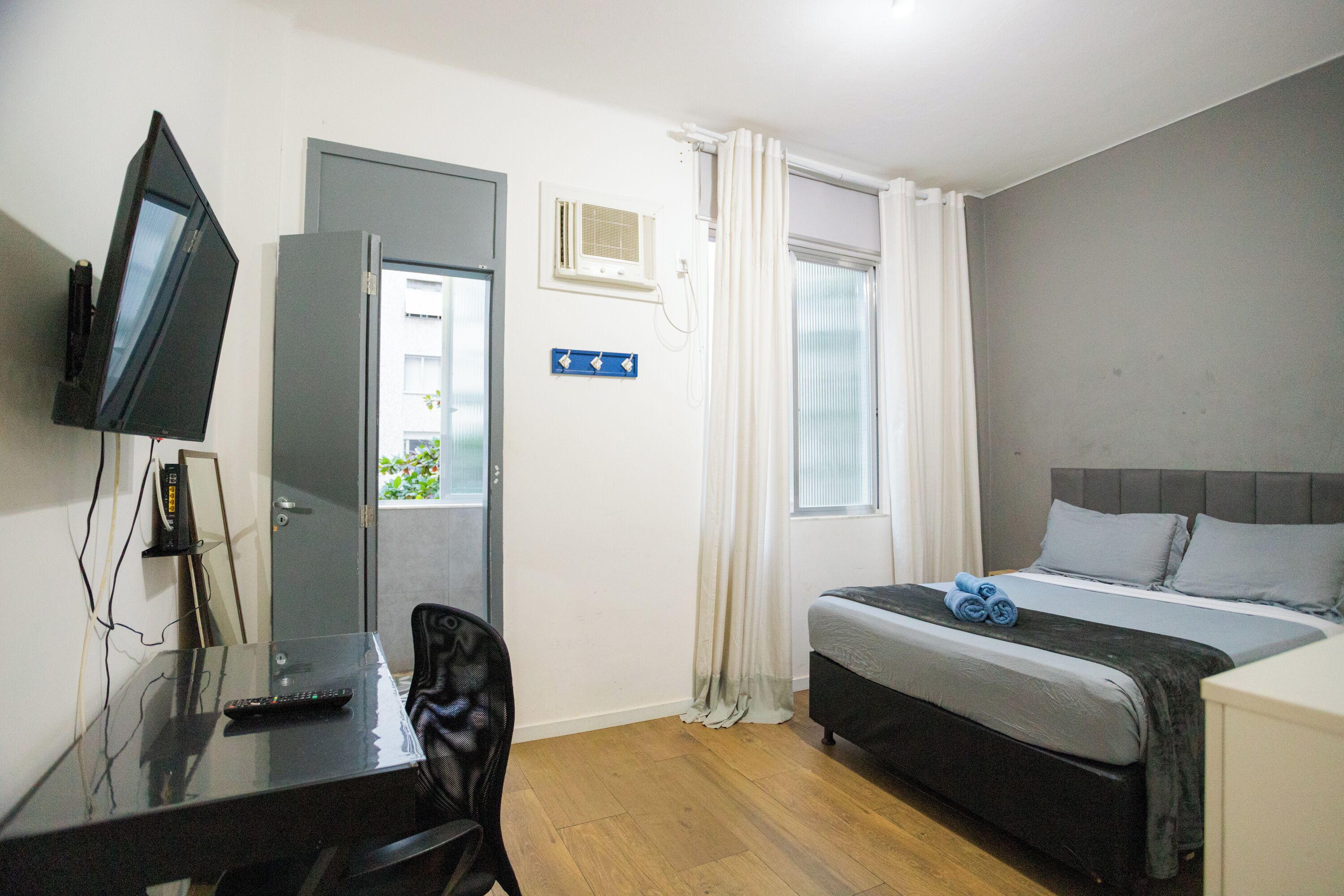 A hotel room with a bed, a desk with a TV, a chair, and a window with curtains.