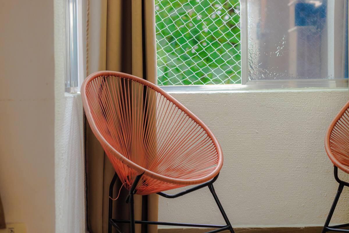 Dos sillas de alambre color naranja ubicadas cerca de una ventana con mosquitero verde.