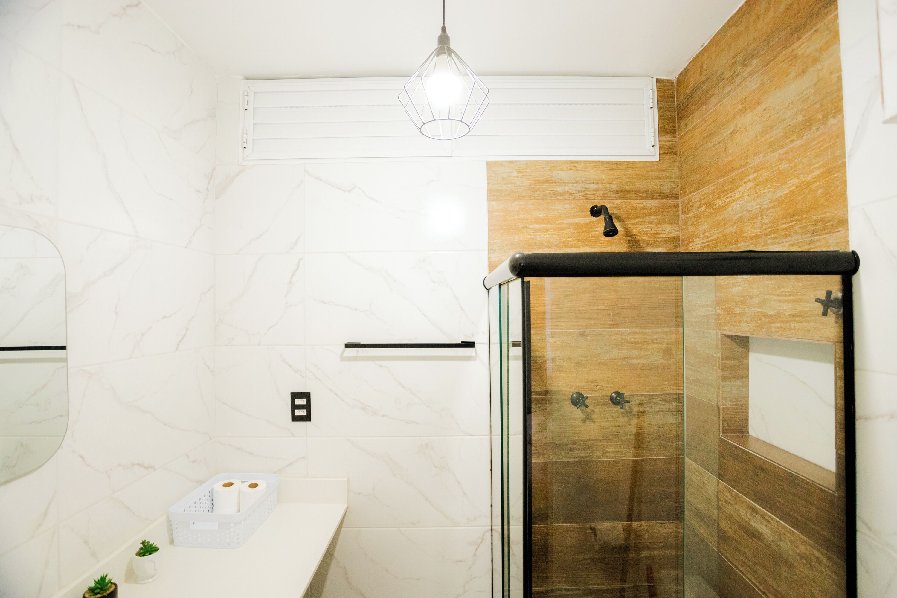 A modern bathroom with a marble wall, a wooden accent wall, a glass shower enclosure, and a white countertop with a small plant.