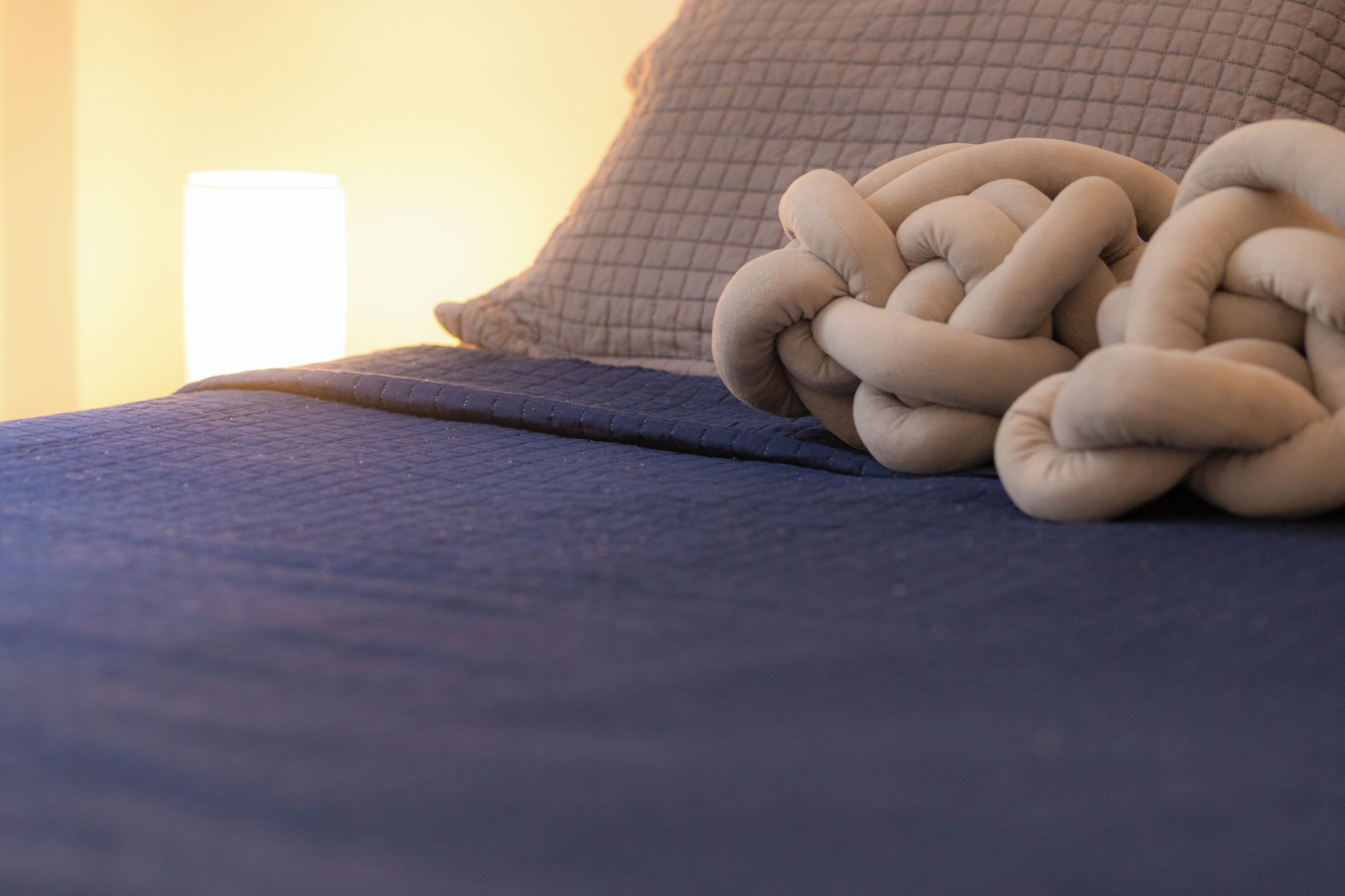 A bed with a blue cover and a beige, knotted throw blanket.