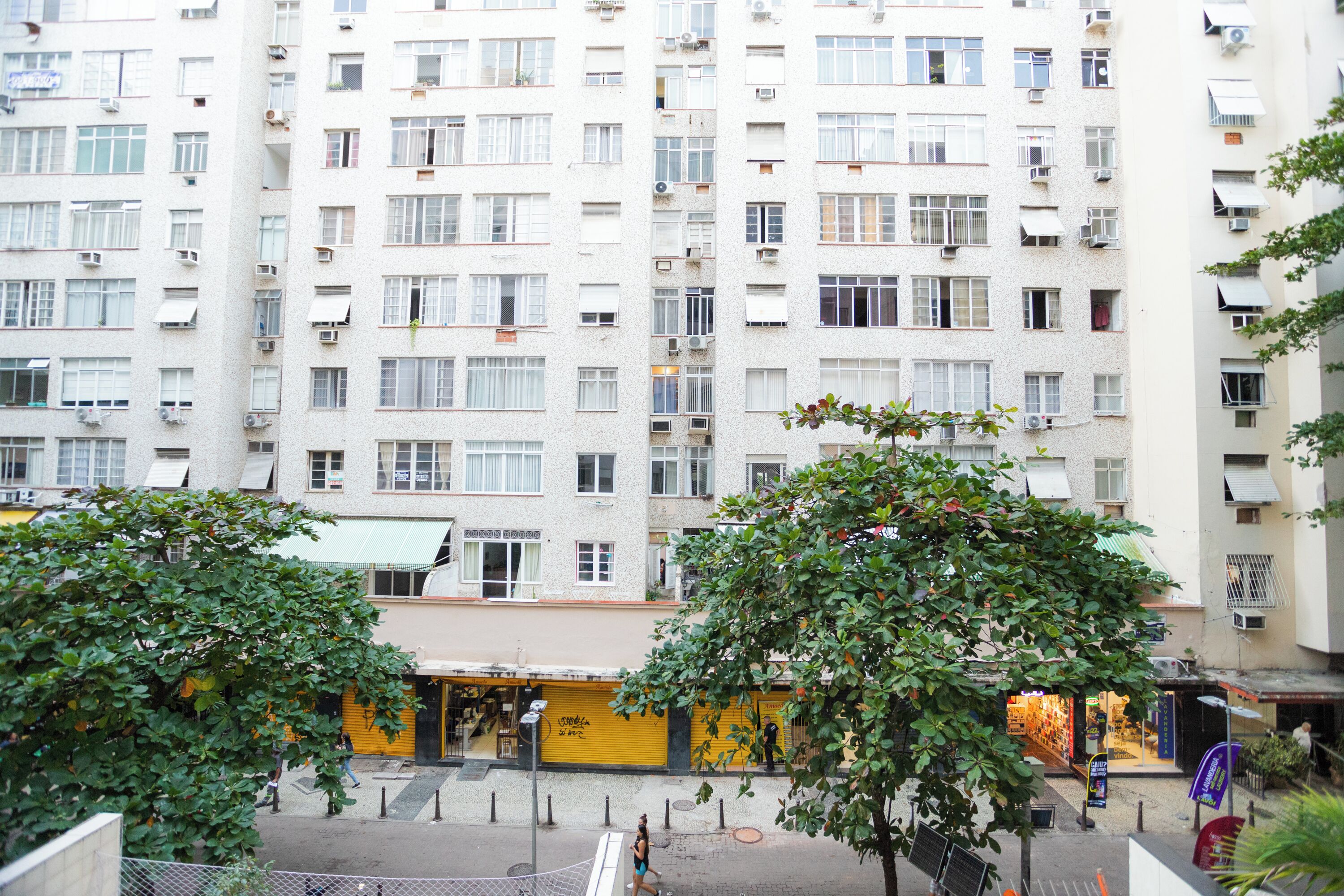 A multi-story residential building with balconies, a yellow storefront, and trees in the foreground.