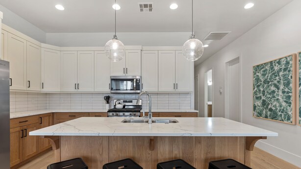 Cocina moderna con gabinetes blancos, una isla de mármol, taburetes negros e iluminación colgante.