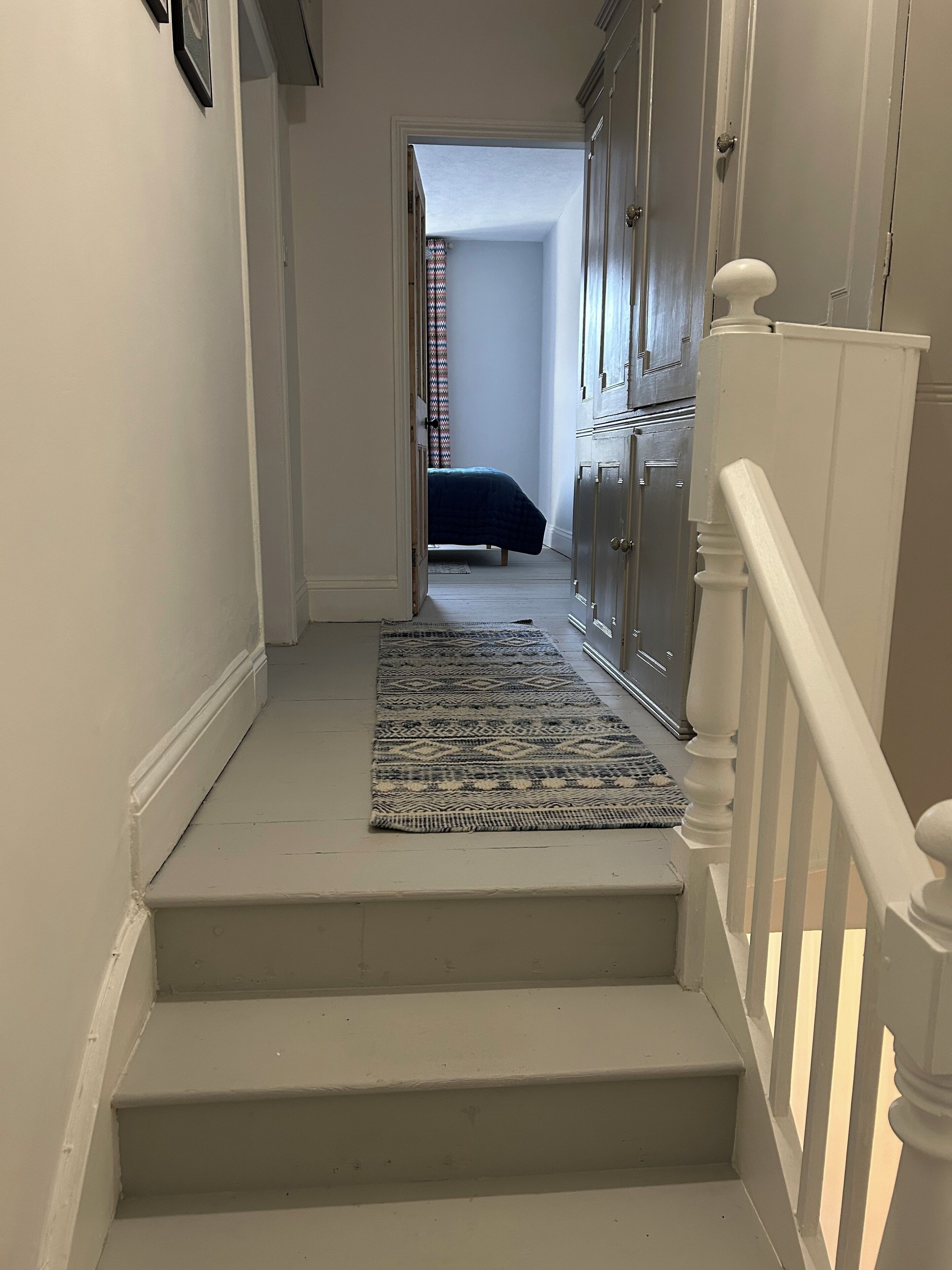 Una escalera conduce a un dormitorio con cama, cómoda y ventana con persianas.
