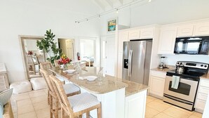 A modern kitchen with a central island, white cabinetry, stainless steel appliances, and a dining area with wicker chairs. - Experience the exquisite Mango Serenity, a spacious condo that offers breathtaking views and an abundance of comfort.  Discover your perfect getaway today! (St John)