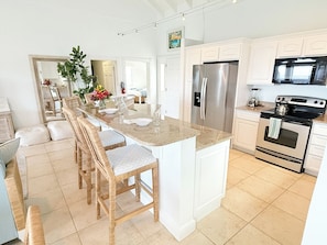 A modern kitchen with a central island, white cabinetry, stainless steel appliances, and a dining area with wicker chairs. - Experience the exquisite Mango Serenity, a spacious condo that offers breathtaking views and an abundance of comfort.  Discover your perfect getaway today! (St John)