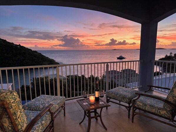 A balcony with two patterned chairs, a table with a candle and wine glasses, and a view of the sea at sunset.
