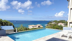 A swimming pool with a view of the ocean, white lounge chairs, and a building with a blue roof. - Experience the exquisite Mango Serenity, a spacious condo that offers breathtaking views and an abundance of comfort.  Discover your perfect getaway today! (St John)