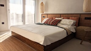 A neatly made bed with a wooden frame and white bedding, a woven headboard, and a small round ottoman beside it.