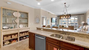 A kitchen with granite countertops, a stainless steel sink, and wooden cabinets. There is a built-in shelving unit with decorative plates and a small fish tank. - Adagio G-403 Poolside 3/3 - NEW to program for 2025! (Santa Rosa Beach)