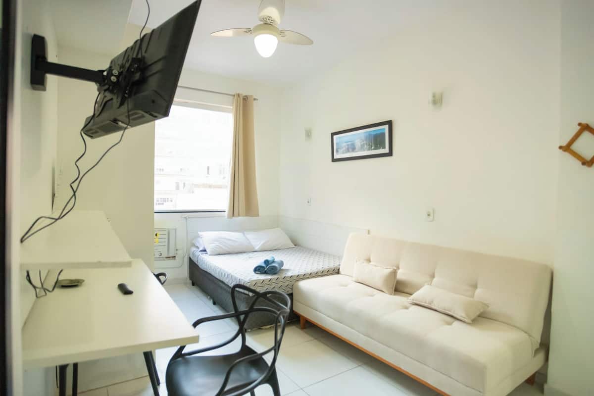 A small, well-lit room with a bed, a sofa, a desk, and a TV mounted on the wall.