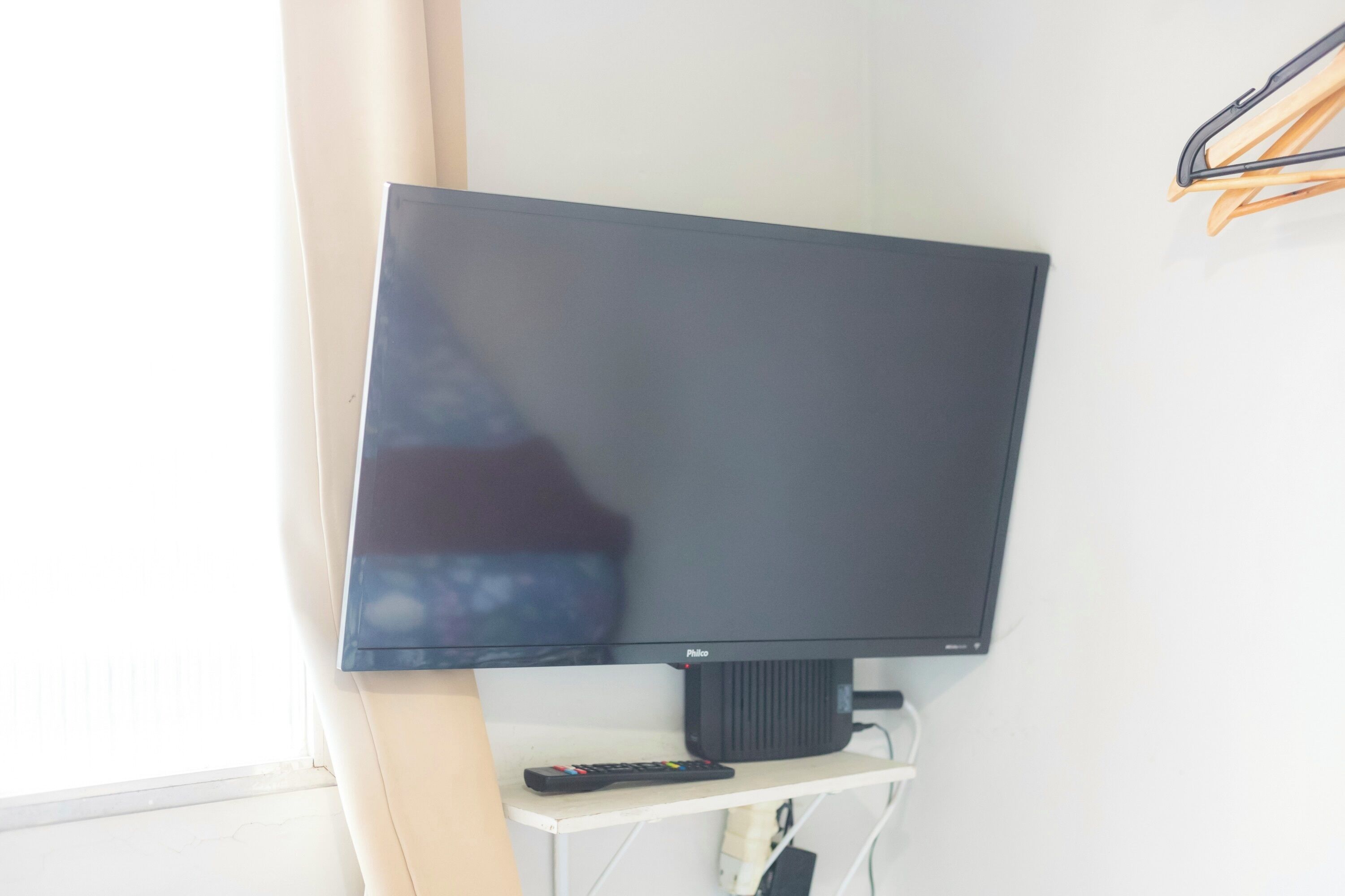 A flat-screen TV mounted on a wall, a remote control on a stand below it, and a power outlet to the right.