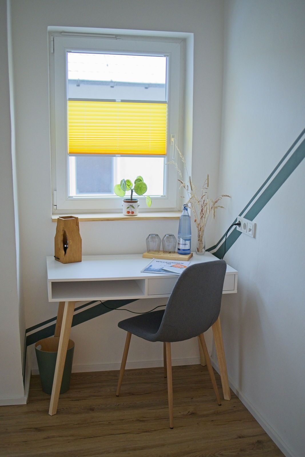 Ein kleiner Arbeitsbereich mit Schreibtisch, Stuhl, Fenster mit Jalousien und einer Topfpflanze.