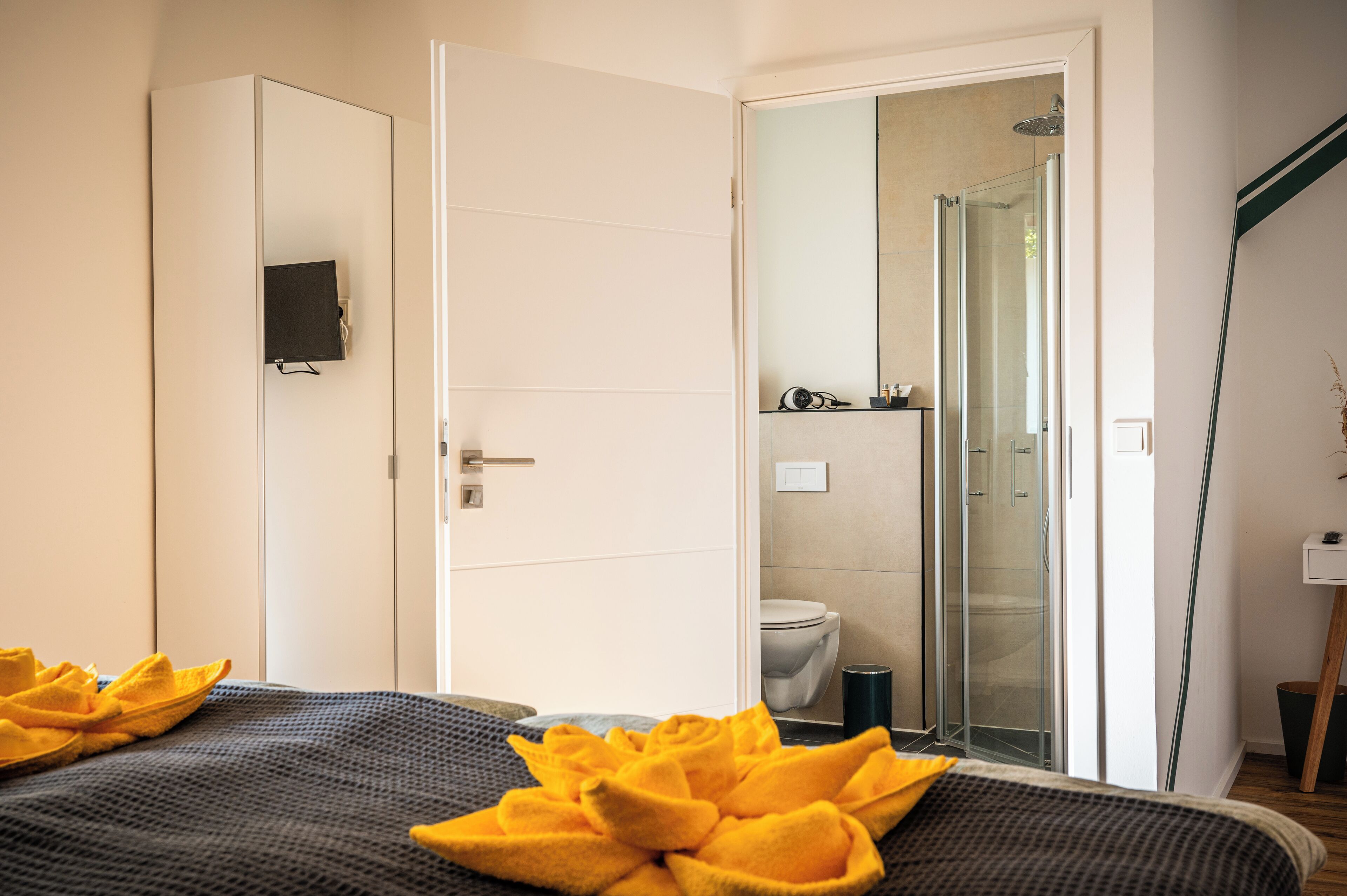Quarto de hotel moderno com uma cama, área de chuveiro e vaso sanitário visíveis através de uma porta de vidro.