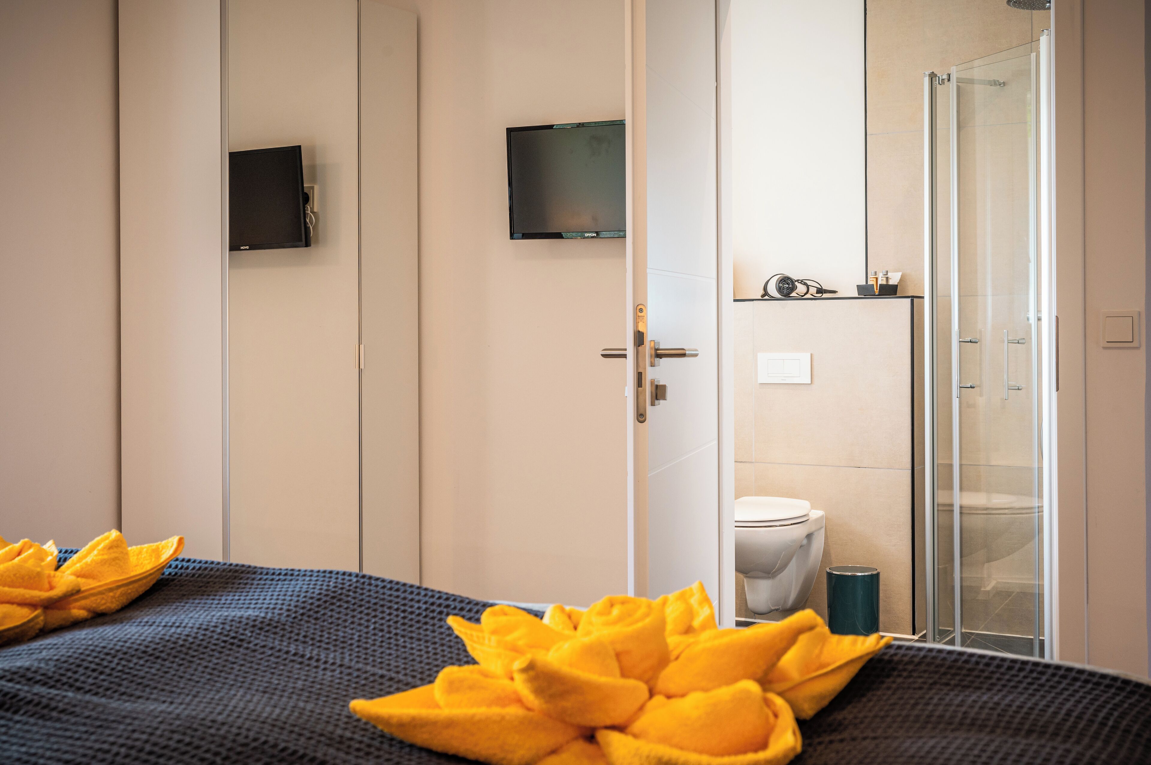 Habitación de hotel con cama, ducha, inodoro y televisor montado en la pared.