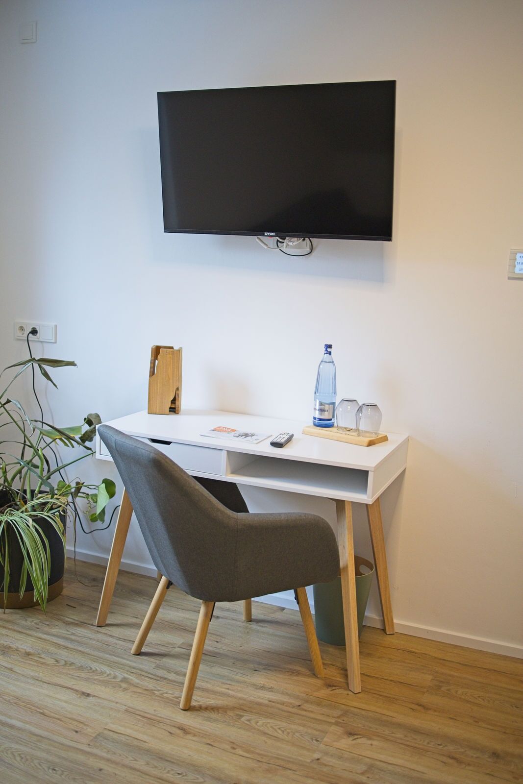 A room with a white desk, a grey chair, a television mounted on the wall, a potted plant, and a bottle of water.