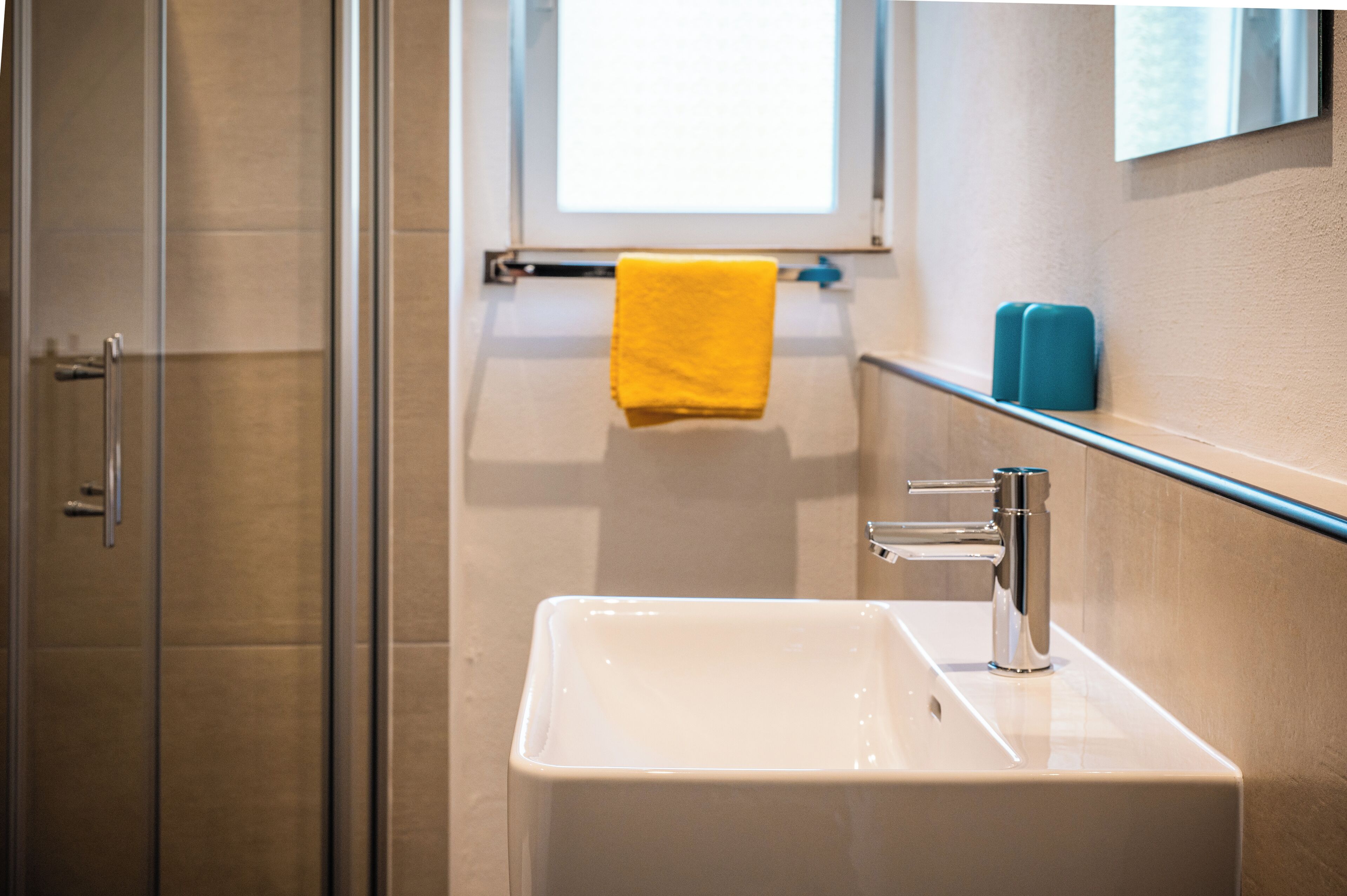 A bathroom with a glass shower enclosure, a white sink with a chrome faucet, a yellow towel, and a blue soap dispenser.