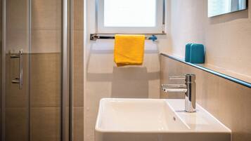 Une salle de bain avec une douche à parois de verre, un lavabo blanc avec un robinet chromé, une serviette jaune et un distributeur de savon bleu.