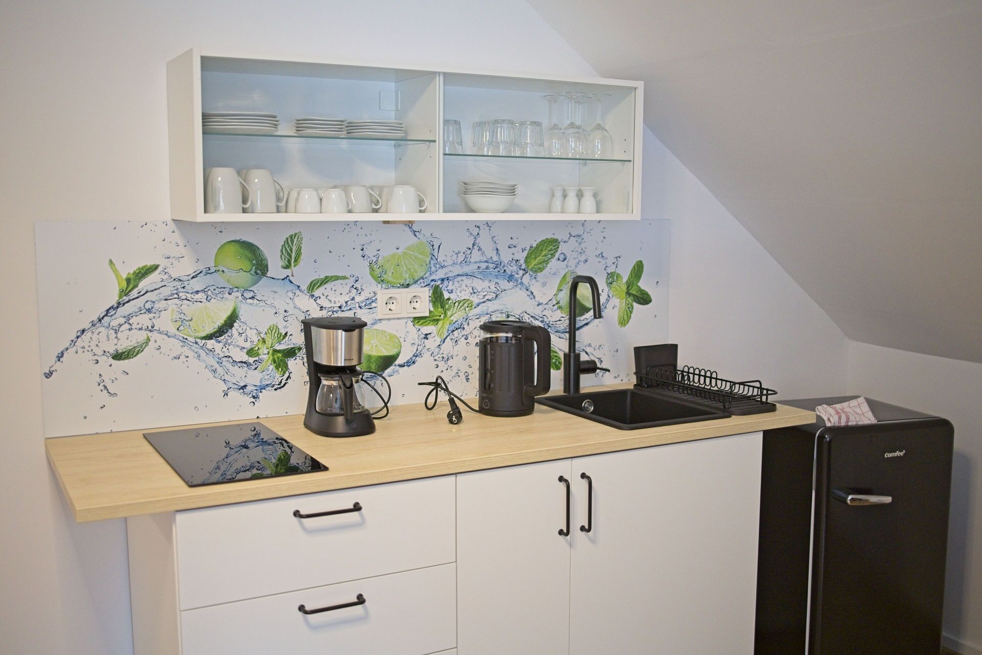 Una cocina con cafetera, lavamanos y refrigerador.