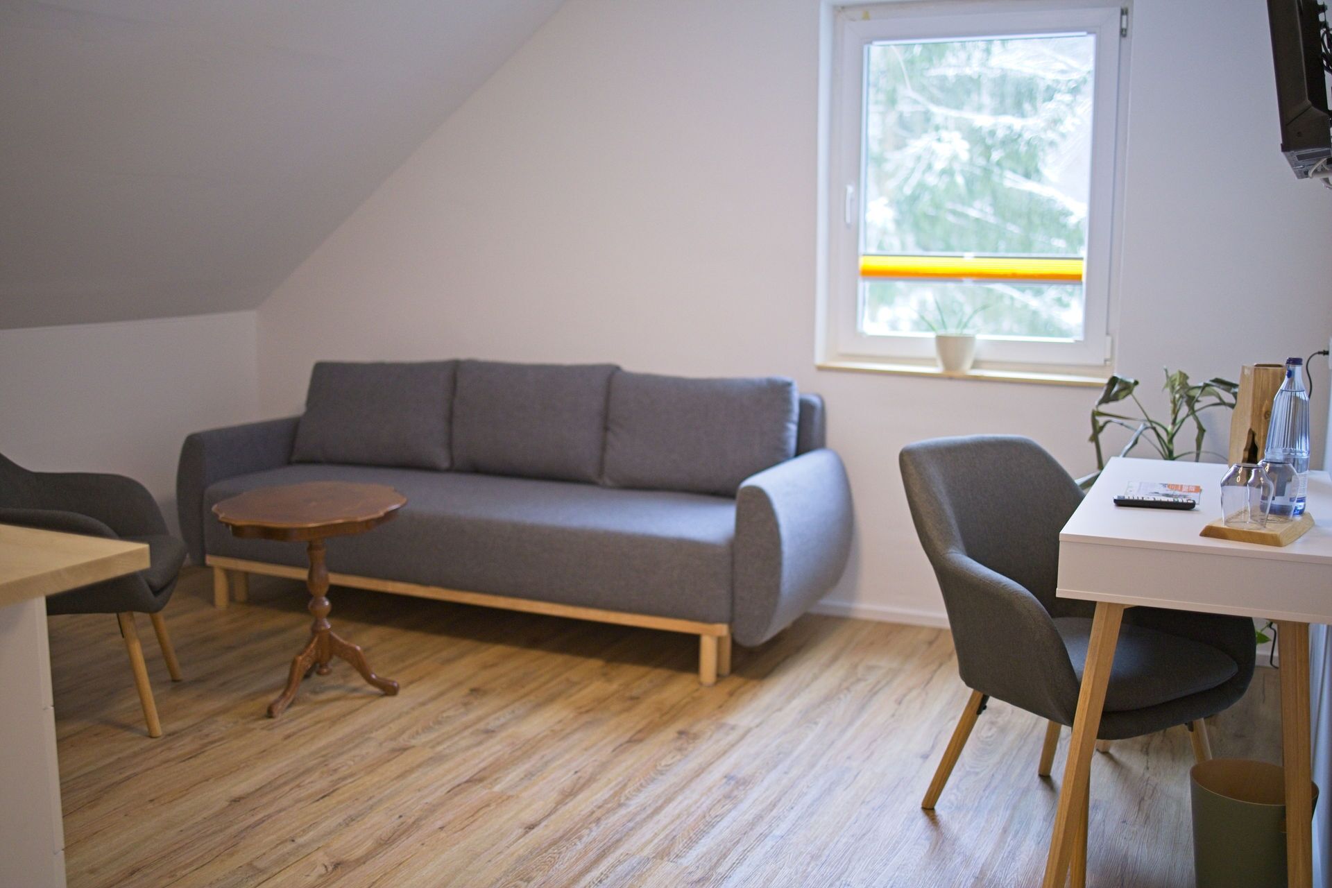 A modern living room with a grey sofa, a small round table, and a desk with a chair. There is a window with a view of trees and a plant on the windowsill.