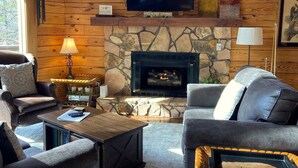 A cozy living room with a stone fireplace, leather sofas, a television mounted above the fireplace, and a mounted artwork on the wall. - Sugar Maple Sunset in Shagbark (Sevierville)