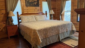 A wooden cabin bedroom with a bed, nightstands, lamps, and a window with curtains. - Sugar Maple Sunset in Shagbark (Sevierville)
