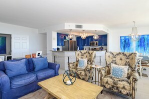 A modern living room with a blue sofa, patterned armchairs, a coffee table, and a kitchen in the background. - Coastal Beachfront Condo w/ Beach Service Included in Destin! 🏖🐚 (Destin)