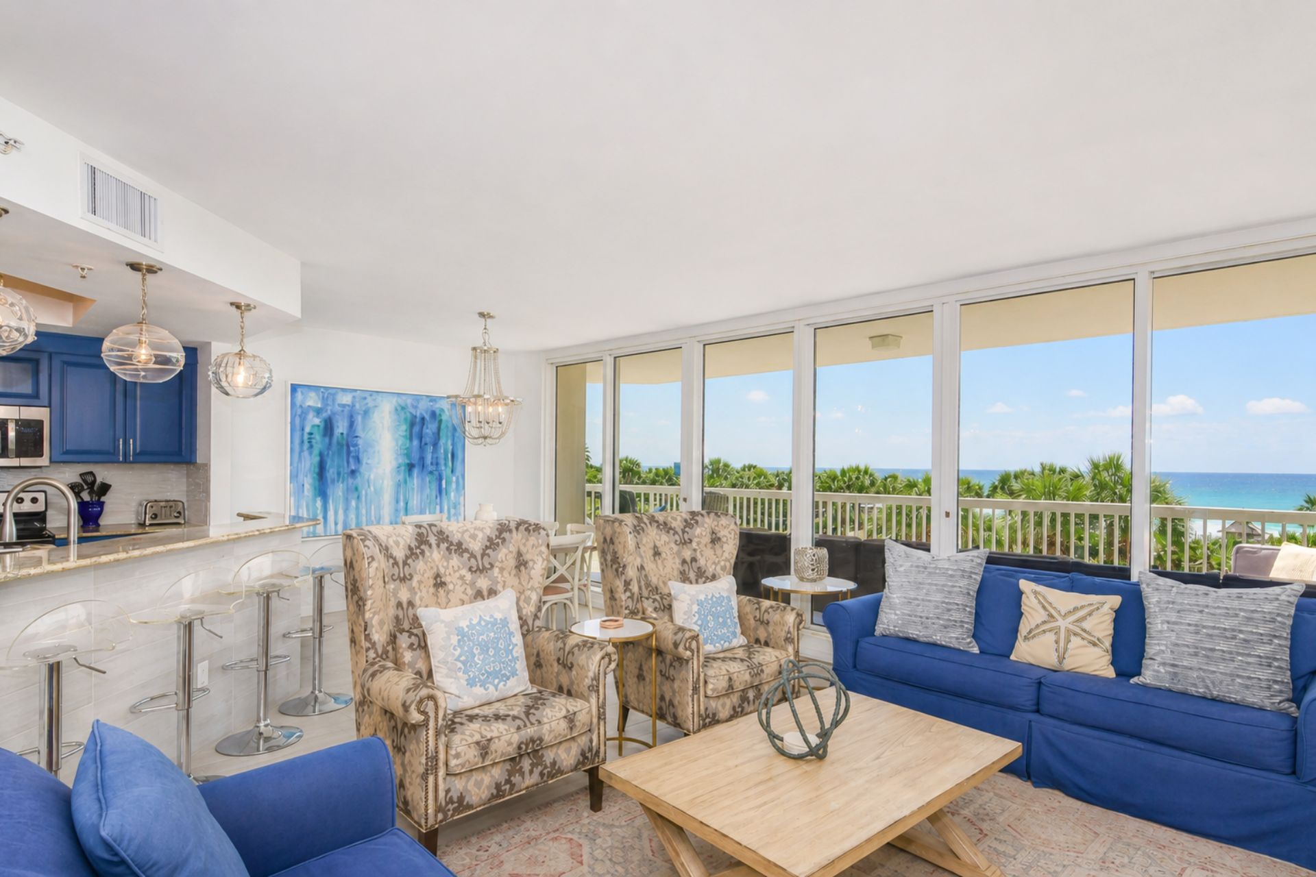 A spacious living area with a large window overlooking the ocean, a kitchen with blue cabinets, and a balcony with outdoor seating.