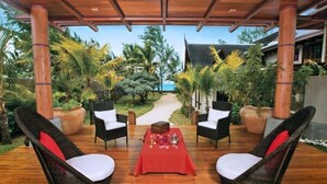 Un espace extérieur abrité, aménagé avec des chaises en osier et une table rouge, entouré de plantes tropicales et bordé d’un chemin menant à un bâtiment.