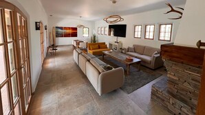 A spacious living room with a sectional sofa, a coffee table, and a TV. There are multiple windows providing natural light.