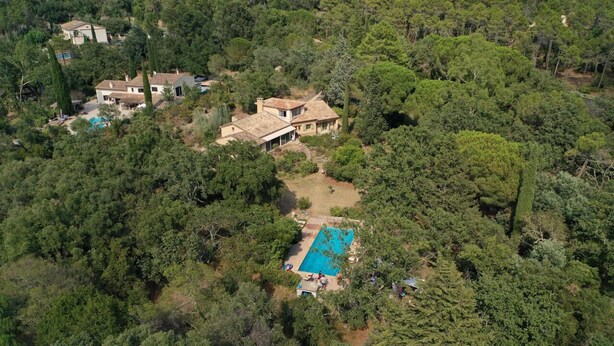 An aerial view of a property with a large house surrounded by dense forest, a swimming pool, and a small garden.