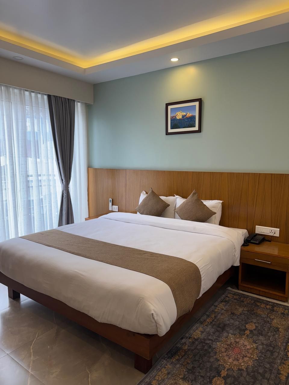 Habitación de hotel con una cama grande, una mesita de noche, un teléfono, un cuadro en la pared y una ventana con cortinas.