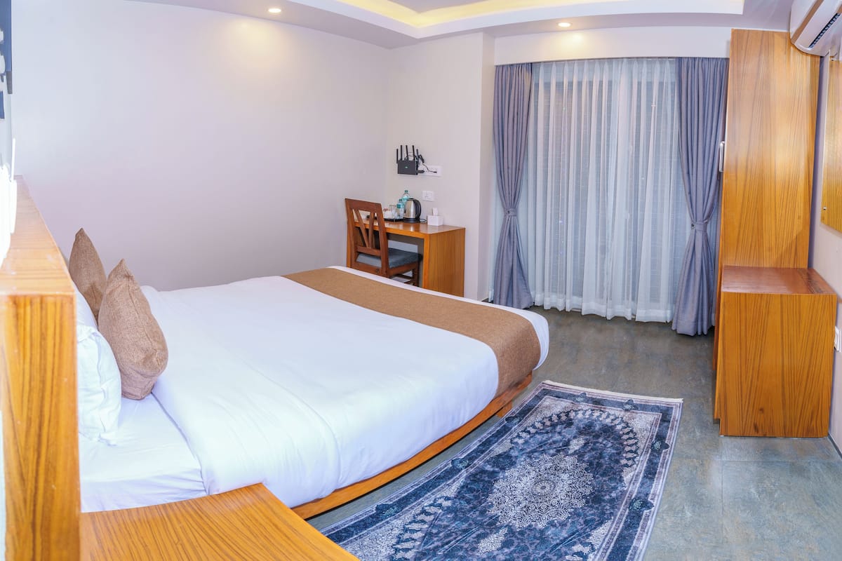 A modern hotel room with a large bed, a wooden desk, a chair, a small table, and a large window with curtains.