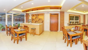 A spacious dining area with multiple wooden tables and chairs, a central counter, and large windows providing natural light. - Bay Hotel (Pokhara)