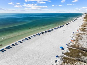 An aerial view of a beach with rows of blue umbrellas and beach chairs, a sandy shoreline, and a clear blue ocean.