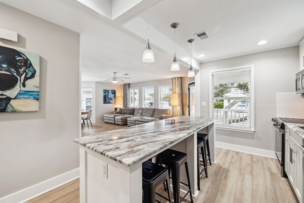 A modern kitchen with a marble countertop, black bar stools, and a large window with blinds.